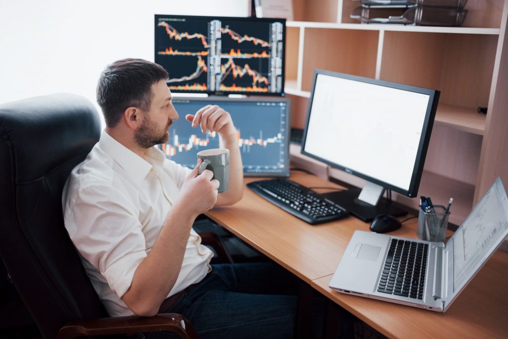 Young businessman is sitting in office at table, working on computer with many monitors,diagrams on